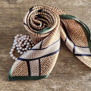 May include: A rolled silk scarf with a geometric pattern in tan, black, and gold. The scarf has a dark green border and is paired with a strand of pink pearls. The scarf is displayed on a wooden surface.