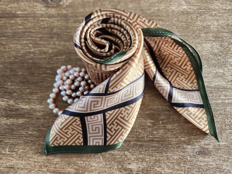 May include: A rolled silk scarf with a geometric pattern in tan, black, and gold. The scarf has a dark green border and is paired with a strand of pink pearls. The scarf is displayed on a wooden surface.