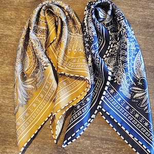 May include: Two silk bandanas, one mustard yellow and one navy blue, are displayed. Both feature intricate paisley and feather patterns with white borders. The bandanas are folded, showcasing their detailed designs and textures.