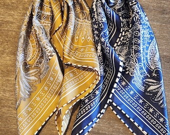 May include: Two silk bandanas, one mustard yellow and one navy blue, are displayed. Both feature intricate paisley and feather patterns with white borders. The bandanas are folded, showcasing their detailed designs and textures.