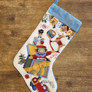 May include: A white Christmas stocking with a blue cuff and hanging loop. The stocking features cross-stitch images of a toy soldier, a clown, a reindeer, and a toy train. The images are in red, blue, green, yellow, and brown.