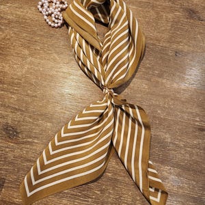 May include: A mustard-coloured silk scarf with white stripes, knotted at the centre. A strand of faux pearls rests beside the scarf. The scarf's design features alternating stripes, creating a chevron pattern at the ends. The scarf is a fashion accessory.