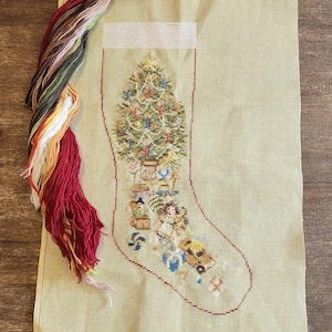 May include: A Christmas stocking needlepoint canvas with a festive tree design, featuring ornaments and gifts. The canvas is light green with a red outline, and colourful embroidery floss is included.