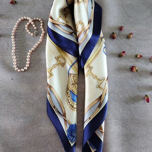 May include: Ivory and navy blue silk scarf with a decorative equestrian-themed pattern. The scarf features gold, blue, and brown accents, and is paired with a pearl necklace and dried rosebuds on a gray surface.