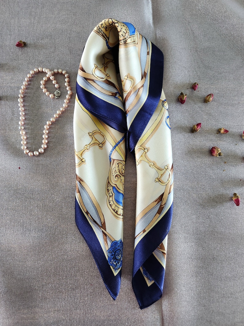 May include: Ivory and navy blue silk scarf with a decorative equestrian-themed pattern. The scarf features gold, blue, and brown accents, and is paired with a pearl necklace and dried rosebuds on a gray surface.