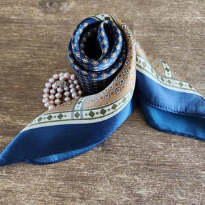 May include: A rolled silk scarf with a geometric pattern in navy blue, tan, and white. The scarf is displayed on a wooden surface, with a pearl necklace next to it. The scarf's design includes small diamond shapes and a border.