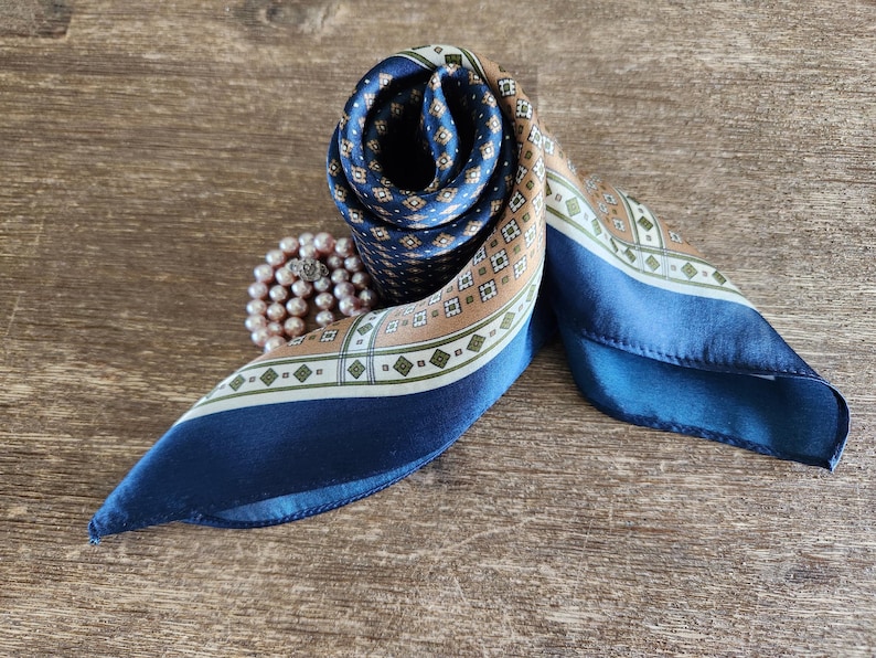 May include: A rolled silk scarf with a geometric pattern in navy blue, tan, and white. The scarf is displayed on a wooden surface, with a pearl necklace next to it. The scarf's design includes small diamond shapes and a border.