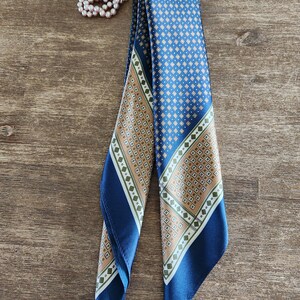 May include: A blue silk scarf with a geometric pattern in tan and white. The scarf is folded and draped, with a matching pearl bracelet in the background. The scarf has a dark blue border.