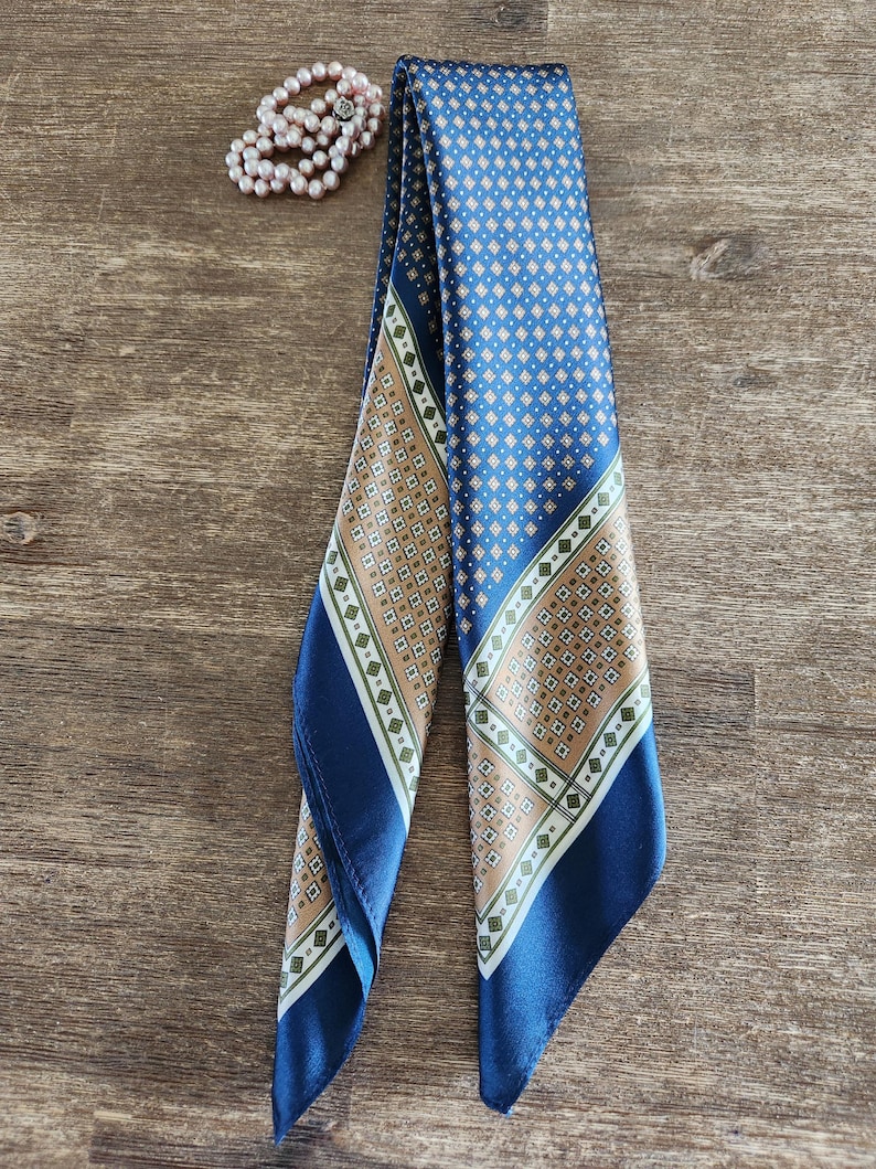 May include: A blue silk scarf with a geometric pattern in tan and white. The scarf is folded and draped, with a matching pearl bracelet in the background. The scarf has a dark blue border.