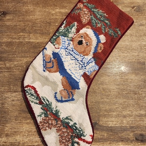 May include: A Christmas stocking with a red top and trim, depicting a bear in a blue and white outfit and ice skates. The bear is surrounded by pine branches and pine cones on a white background.