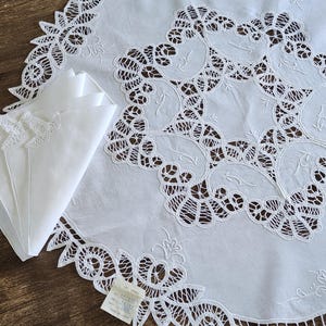 Puede incluir: Mantel de mesa de algodón blanco con un delicado diseño de encaje. El mantel tiene forma circular y presenta un motivo central de intrincados patrones florales. El encaje es blanco y tiene una textura delicada y aireada.