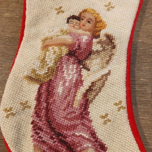 May include: A Christmas stocking with a detailed needlepoint design. The stocking features a red border and a cream-colored background. The needlepoint depicts an angel holding a child, with gold accents and star patterns.
