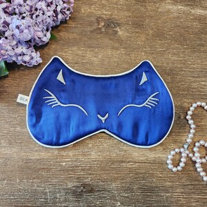 May include: A blue silk sleep mask with a white cat face design. The mask has a white trim and the word "SILK" is printed on the tag.