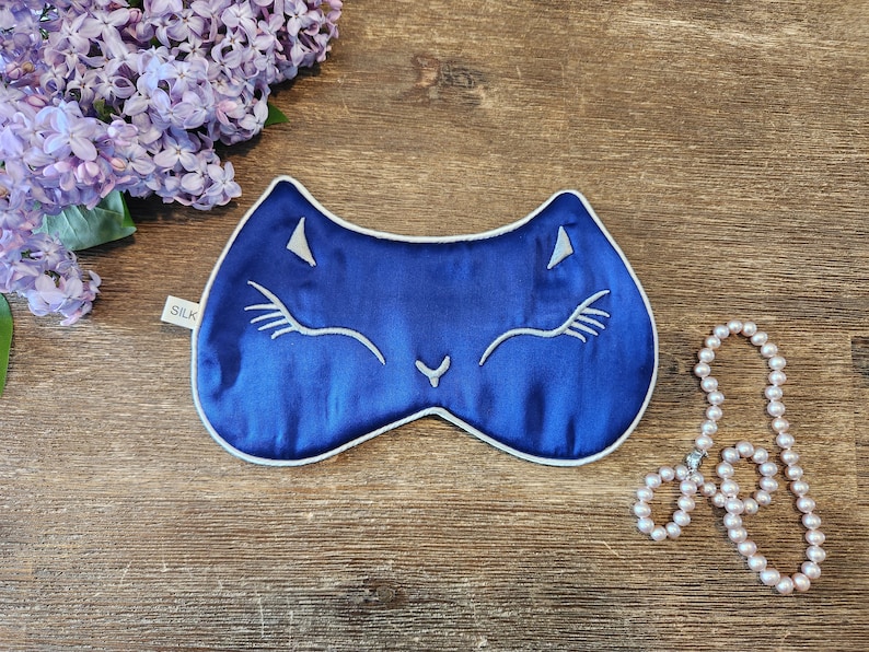 May include: A blue silk sleep mask with a white cat face design. The mask has a white trim and the word "SILK" is printed on the tag.