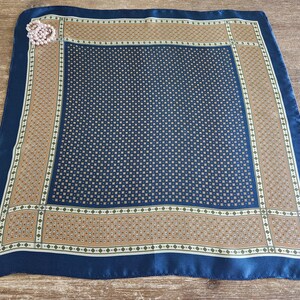May include: A square silk scarf with a navy blue border and a central pattern of small squares. The scarf has a tan and white patterned border. A pink brooch is pinned to the corner of the scarf.