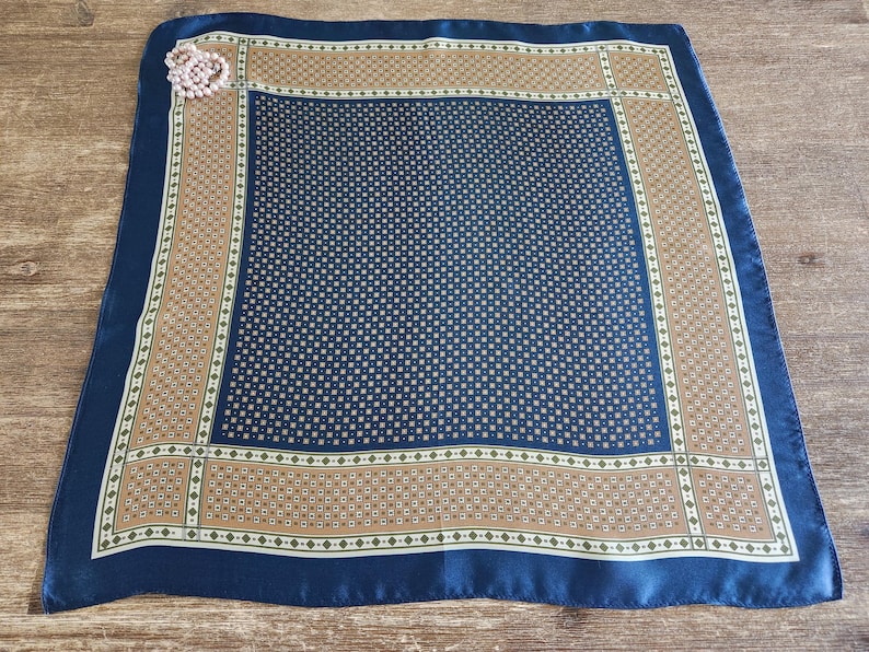 May include: A square silk scarf with a navy blue border and a central pattern of small squares. The scarf has a tan and white patterned border. A pink brooch is pinned to the corner of the scarf.