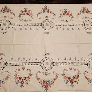 Puede incluir: Mantel blanco con diseños florales bordados y ribete de encaje. El mantel presenta un patrón repetitivo de guirnaldas florales y bordes de encaje delicados.