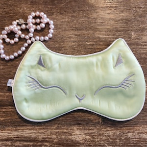 May include: A light green sleep mask with a white trim and a cute embroidered cat face. The cat has closed eyes and a small nose.
