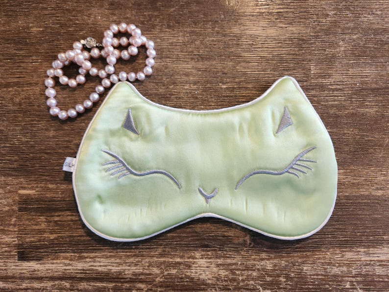 May include: A light green sleep mask with a white trim and a cute embroidered cat face. The cat has closed eyes and a small nose.