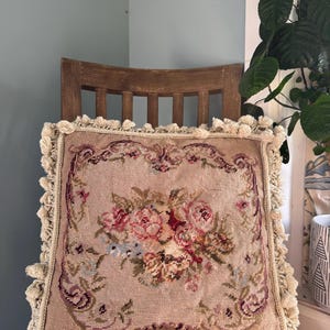 May include: Square, beige needlepoint cushion featuring a floral design in pink, red, and blue hues. The cushion has a cream-coloured fringe trim and is placed on a wooden chair. The floral pattern is centrally positioned.