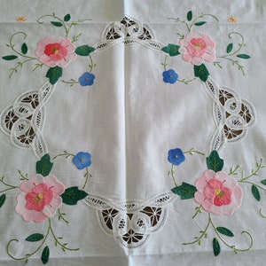 May include: White linen tablecloth with embroidered floral design. Features pink and red roses, blue flowers, and green leaves. The centre has a decorative lace border. A delicate and elegant piece.