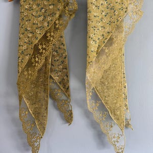 May include: Two triangular scarves in a muted olive green fabric featuring a small white and dark green floral pattern. Each scarf is finished with a wide, intricate scalloped lace border in a matching olive green tone, hanging from a wooden hanger.