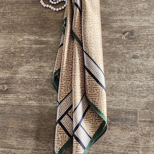 May include: A folded silk scarf with a geometric pattern in shades of tan, black, and white, with a dark green border. A pearl necklace is draped nearby. The scarf is likely a fashion accessory.