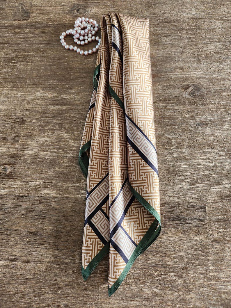 May include: A folded silk scarf with a geometric pattern in shades of tan, black, and white, with a dark green border. A pearl necklace is draped nearby. The scarf is likely a fashion accessory.