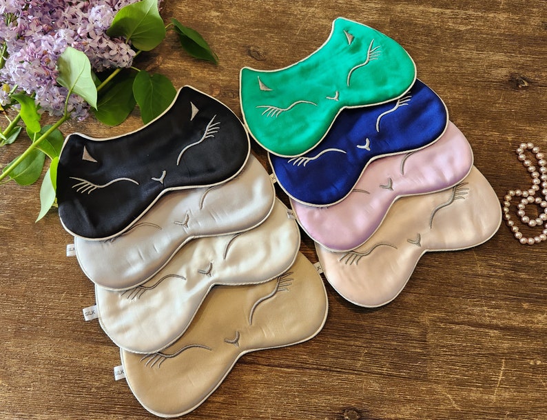 May include: A stack of six sleep masks featuring a cat face design. The masks are made from silk and come in a range of colours, including black, green, blue, pink, and beige.