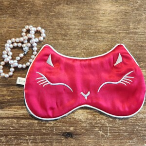 May include: A red satin sleep mask with a white embroidered cat face. The mask has a white trim around the edges.