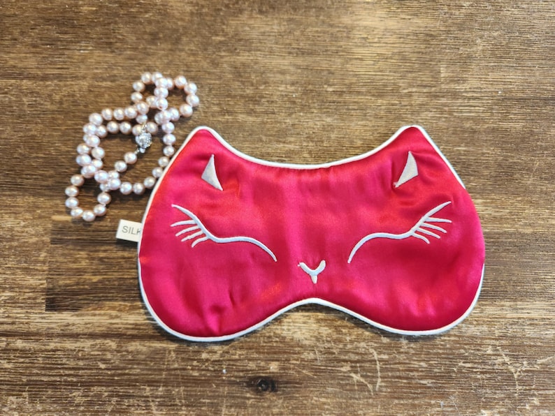 May include: A red satin sleep mask with a white embroidered cat face. The mask has a white trim around the edges.