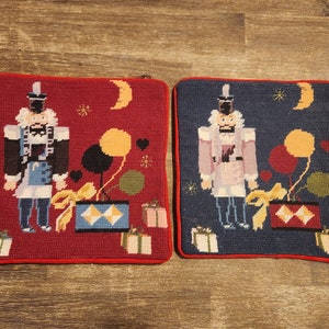 Op de afbeelding: Twee vierkante kussens in rood en blauw met een kerstthema. Elk kussen heeft een notenkraker die een ballon en een cadeaudoos met strik vasthoudt. De kussens hebben een rode en blauwe rand.