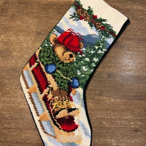 May include: Needlepoint Christmas stocking featuring a teddy bear in a red hat and green jacket sledding down a snowy hill. The stocking has a cream-colored background with a black trim and a loop for hanging. Festive holiday decor.