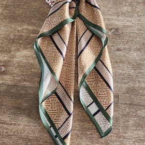 May include: A tan and black patterned silk scarf with a green border. The scarf is folded and tied, with a pearl necklace draped over it. The pattern is geometric and the scarf is on a wooden surface.