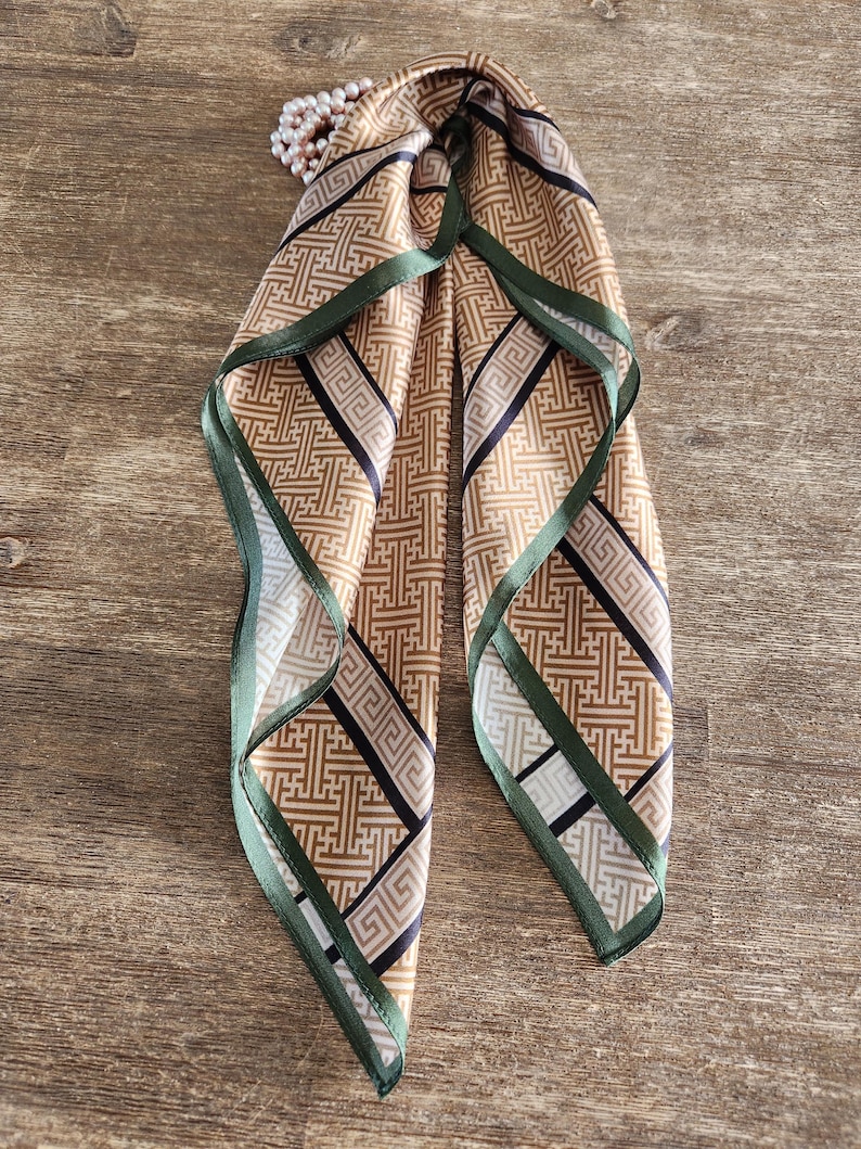 May include: A tan and black patterned silk scarf with a green border. The scarf is folded and tied, with a pearl necklace draped over it. The pattern is geometric and the scarf is on a wooden surface.