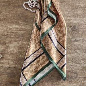 May include: A folded silk scarf with a geometric pattern in shades of tan, black, and green. The scarf has a repeating design and is trimmed with a green border. A pearl necklace is next to the scarf on a wooden surface.