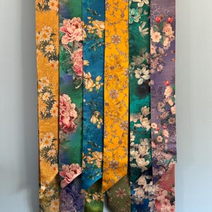 May include: A collection of seven silk scarves with floral patterns in various colors, including yellow, green, blue, and purple. Each scarf is vertically displayed on a wooden hanger, showcasing the intricate designs and vibrant hues.