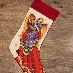 Puede incluir: Una media de Navidad de terciopelo rojo con un diseño de punto de aguja de un ángel tocando un arpa. El ángel lleva una túnica morada con ribetes dorados y un vestido rojo. La media está decorada con estrellas doradas.