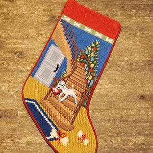 May include: Needlepoint Christmas stocking with a red cuff and trim. The stocking features a scene of a white dog carrying a heart on a staircase next to a Christmas tree. The background includes a blue sky and a white door.