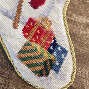 K&ouml;nnte beinhalten: Ein wei&szlig;er Weihnachtssocken mit goldener Kordel. Der Socken ist mit einer Winterszene bestickt, die einen Schneemann, Geschenke und einen Baum zeigt. Die Geschenke sind in rotes, gr&uuml;nes und blaues Papier eingepackt.