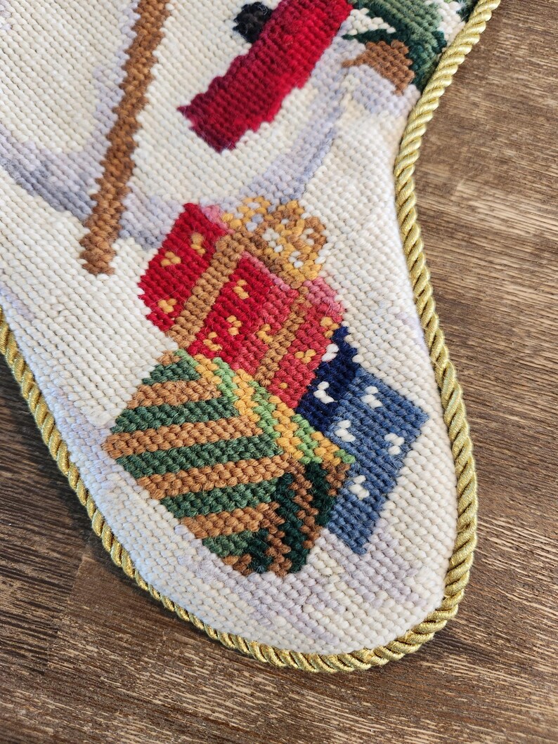 K&ouml;nnte beinhalten: Ein wei&szlig;er Weihnachtssocken mit goldener Kordel. Der Socken ist mit einer Winterszene bestickt, die einen Schneemann, Geschenke und einen Baum zeigt. Die Geschenke sind in rotes, gr&uuml;nes und blaues Papier eingepackt.