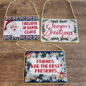 May include: Three rectangular Christmas ornaments with gold rope hangers. One says "I BELIEVE IN SANTA CLAUS" with a Santa Claus image. Another says "Season's Greetings." The third says "FRIENDS ARE THE BEST PRESENTS."