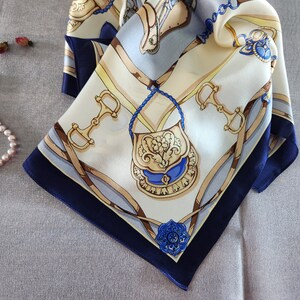 May include: A square silk scarf with a cream background and a navy blue border. The scarf features a detailed equestrian-themed design with gold, blue, and brown accents. The design includes horse bits, a purse, and decorative elements.