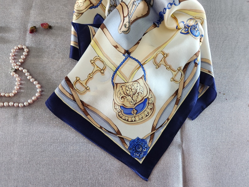 May include: A square silk scarf with a cream background and a navy blue border. The scarf features a detailed equestrian-themed design with gold, blue, and brown accents. The design includes horse bits, a purse, and decorative elements.