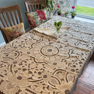Pode incluir: Uma toalha de mesa de renda bege com intrincados padrões florais cobre uma mesa de madeira. Duas almofadas decorativas com desenhos florais repousam sobre cadeiras de madeira. Um pequeno vaso de vidro com vegetação está sobre a mesa.