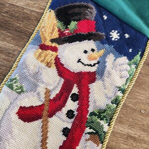K&ouml;nnte beinhalten: Ein wei&szlig;er Weihnachtsstrumpf aus Nadelfilz mit einem Schneemann-Motiv. Der Schneemann tr&auml;gt einen roten Schal und einen schwarzen Zylinder. Der Strumpf hat einen goldenen Besatz und eine gr&uuml;ne Filzmanschette.