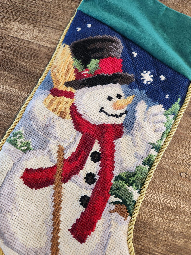K&ouml;nnte beinhalten: Ein wei&szlig;er Weihnachtsstrumpf aus Nadelfilz mit einem Schneemann-Motiv. Der Schneemann tr&auml;gt einen roten Schal und einen schwarzen Zylinder. Der Strumpf hat einen goldenen Besatz und eine gr&uuml;ne Filzmanschette.