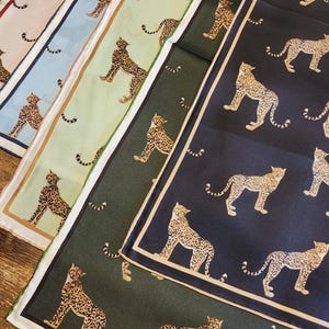 May include: A collection of colorful silk scarves featuring a repeating pattern of a leopard. The scarves are in various colors, including pink, blue, green, and navy, each with a contrasting border. The leopard design is in shades of brown and tan.