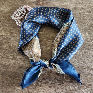 May include: A navy blue silk scarf with a geometric pattern in white, tan, and green. The scarf is knotted and lies on a wooden surface, with a strand of pink pearls in the background. A stylish accessory.