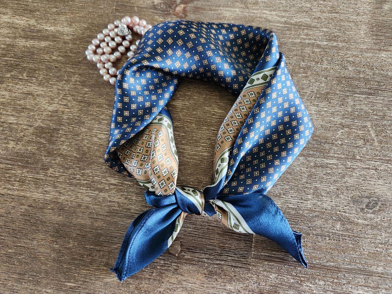 May include: A navy blue silk scarf with a geometric pattern in white, tan, and green. The scarf is knotted and lies on a wooden surface, with a strand of pink pearls in the background. A stylish accessory.